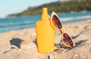 Bir şişe güneş kremi ve kumsalda okyanus kenarında güneş gözlüğü.