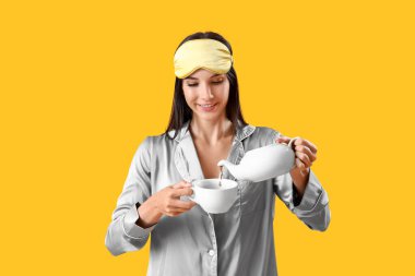 Pijamalı genç bir kadın sarı arka planda fincana çay dolduruyor.