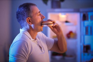 Aç yetişkin bir adam geceleri mutfakta hamburger yiyor, yakın plan.