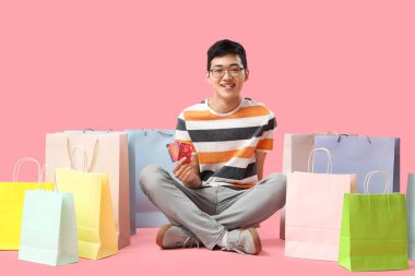 Hediye kartları ve alışveriş torbalarıyla pembe arka planda oturan genç Asyalı adam.