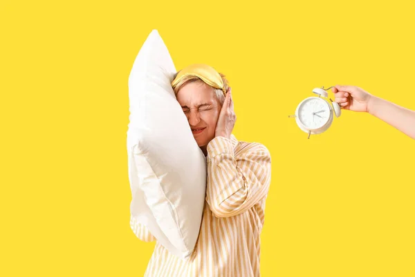 Displeased mature woman in pajamas with pillow and ringing alarm clock on yellow background
