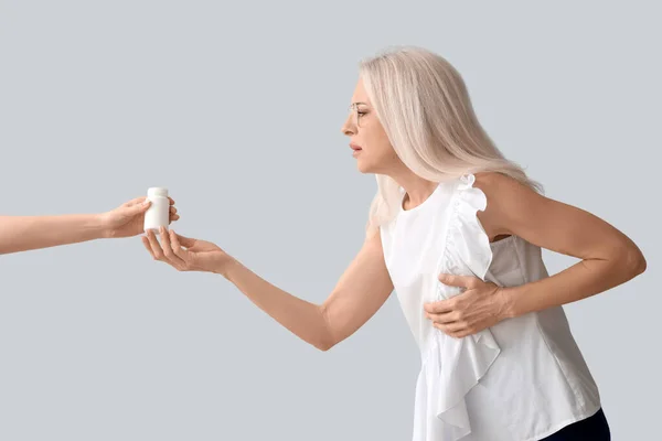 Mature woman with heart attack taking pill bottle on grey background
