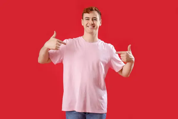 Młody Mężczyzna Wskazuje Swoją Koszulkę Czerwonym Tle — Zdjęcie stockowe
