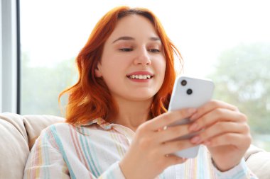 Evdeki koltukta cep telefonu kullanan genç bir kadın.