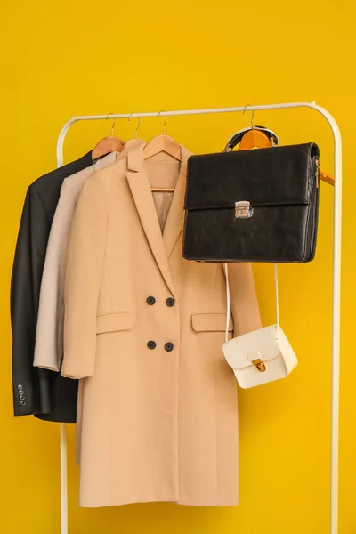 Stylish clothes and bags hanging on rack against yellow background, closeup