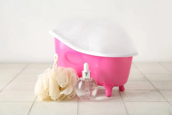 Small bathtub with foam, bottle of essential oil and sponge on light tile table