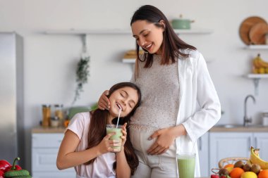 Küçük kız hamile annesiyle mutfakta yeşil smoothie içiyor.