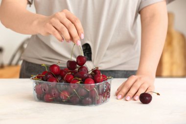 Kadın mutfaktaki masada plastik kutuya olgun kirazlar koyuyor.