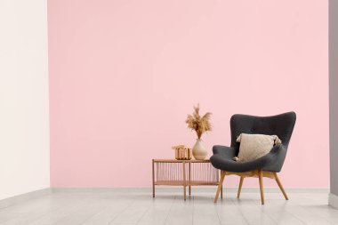 Pembe duvarın yanındaki sehpanın üzerinde pampa otu olan gri koltuk ve vazo.