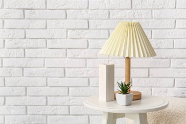Beyaz tuğla duvarın yanında lamba ve çiçek olan küçük bir sehpa.