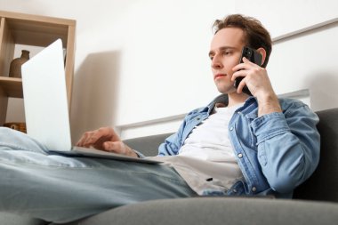 Evde koltukta cep telefonuyla konuşan dizüstü bilgisayarı olan serbest çalışan bir erkek.