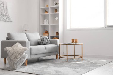 Interior of light living room with grey sofa, table and shelving unit