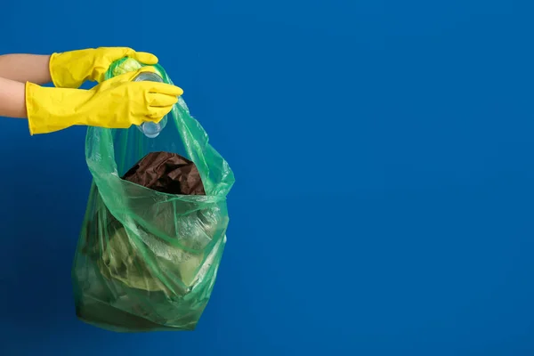 Female hands in rubber gloves putting plastic bottle into garbage bag on blue background