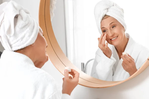 Mature woman applying under-eye cream near mirror in bathroom