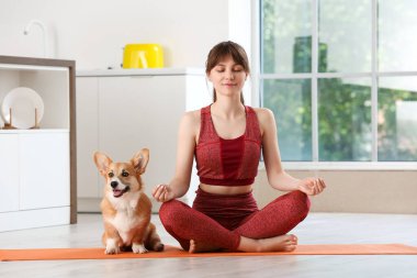 Genç bir kadın, mutfakta yoga minderinde Corgi köpeğiyle meditasyon yapıyor.