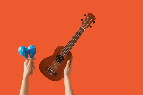 Female hands holding maracas and ukulele on orange background