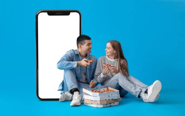 Büyük akıllı telefon ve mutlu genç çift mavi arka planda taze pizza yiyorlar.