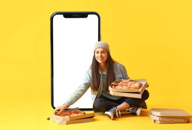 Büyük akıllı telefon ve sarı arka planda bir sürü pizza kutusu olan güzel genç bir kadın.