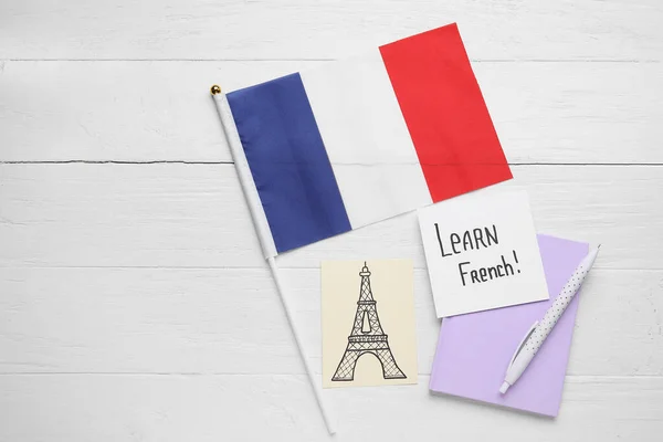Paper with text LEARN FRENCH, drawn Eiffel Tower, notebook and flag on white wooden background