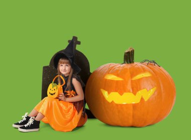 Cadılar Bayramı için cadı gibi giyinmiş şirin küçük kız. Yeşil arka planda büyük bir balkabağı ve mezarı var.