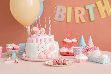 Koyu bej duvarın yanındaki beyaz masada mumlar ve şekerler olan doğum günü pastası.