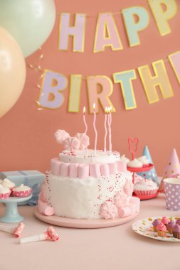 Koyu bej duvarın yanındaki beyaz masada mumlar ve şekerler olan doğum günü pastası.