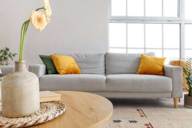 Oturma odasının içinde gri kanepe, kahve masasında gerbera çiçekleri ve büyük pencere.