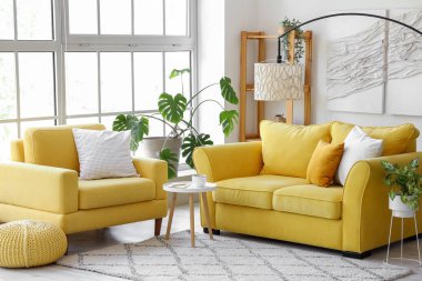 Sarı kanepesi ve büyük pencerenin yanındaki koltuğu olan aydınlık oturma odasının içinde.