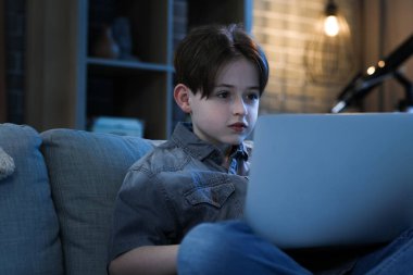 Küçük çocuk dizüstü bilgisayarla ödev yapıyor ve akşamları kanepede oturuyor.