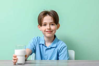 Turkuaz arka planda bir bardak taze sütle masada oturan küçük mutlu çocuk.
