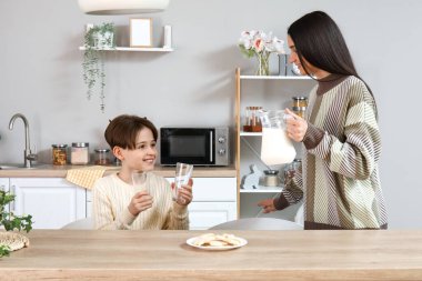 Küçük çocuk bardaklarla masada oturuyor ve annesi mutfakta taze süt dolduruyor.