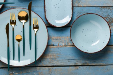 Mavi ahşap masanın üzerinde altın çatal bıçak takımı olan temiz tabaklar.