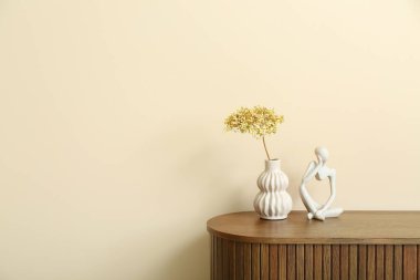 Bej duvarın yanındaki dolapta kurutulmuş ortanca vazosu ve biblo var.