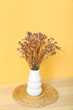 Kurumuş çiçekli vazo ve sarı duvarın yanındaki sehpanın üzerinde paspas.