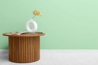 Yeşil duvarın yanındaki sehpanın üzerinde kurutulmuş ortanca vazosu, dergi ve paspas var.