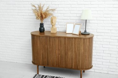 Beyaz tuğla duvarın yanındaki dolapta biblo, fotoğraf çerçevesi ve lamba olan kurutulmuş pampa vazoları.