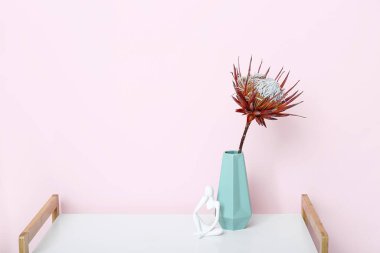 Pembe duvarın yanındaki raf ünitesinde kurumuş kırmızı protea ve biblo olan vazo.