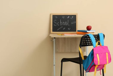 Modern okul masası, sırt çantası, kırtasiye kağıdı ve bej duvarın yanındaki odada Kelime OKULU yazılı karatahta.