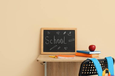 Modern okul masası, sırt çantası, kırtasiye kağıdı ve bej duvarın yanındaki odada Kelime OKULU yazılı karatahta.