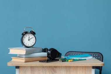 Çalar saati, kulaklığı ve kırtasiyesi olan modern okul masası mavi duvarın yanında.