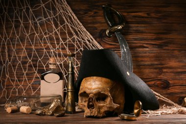 Korsan şapkalı insan kafatası, altın külçeler, kahverengi ahşap arka planda bir şişe rom ve seyahat ekipmanı.