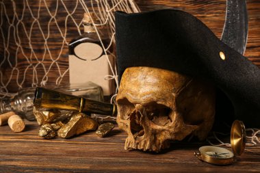 Korsan şapkalı insan kafatası, altın külçeler, kahverengi ahşap arka planda bir şişe rom ve seyahat ekipmanı.