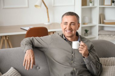 Oturma odasındaki gri koltukta oturan bir fincan kahveyle mutlu olgun bir adam.