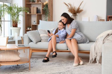 Anne ve kızı cep telefonuyla oturma odasındaki gri kanepede dinleniyorlar.