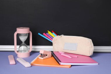 Üzerinde farklı okul kırtasiyesi ve mor masa üzerindeki kum saati olan kalem kutusu.