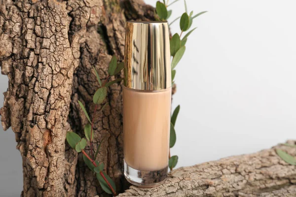 Bottle of makeup foundation, tree bark and eucalyptus branches on light background, closeup