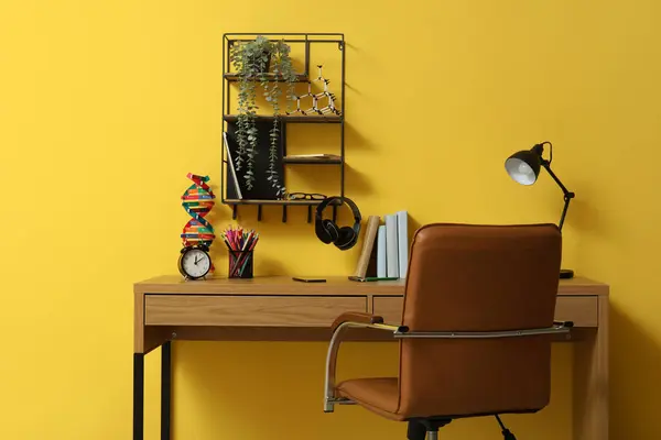 Modern school desk with molecular model, alarm clock and stationery in room near yellow wall