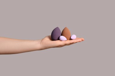 Gri arka planda farklı makyaj süngerleri olan kadın eli