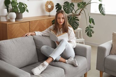 Oturma odasında kanepede kitap okuyan genç bir kadın.