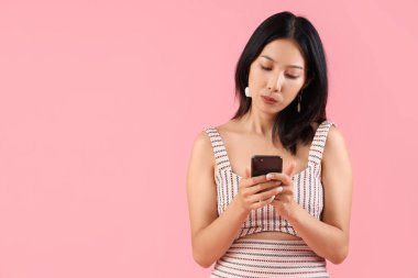 Pembe arka planda cep telefonu olan düşünceli, güzel bir Asyalı kadın.